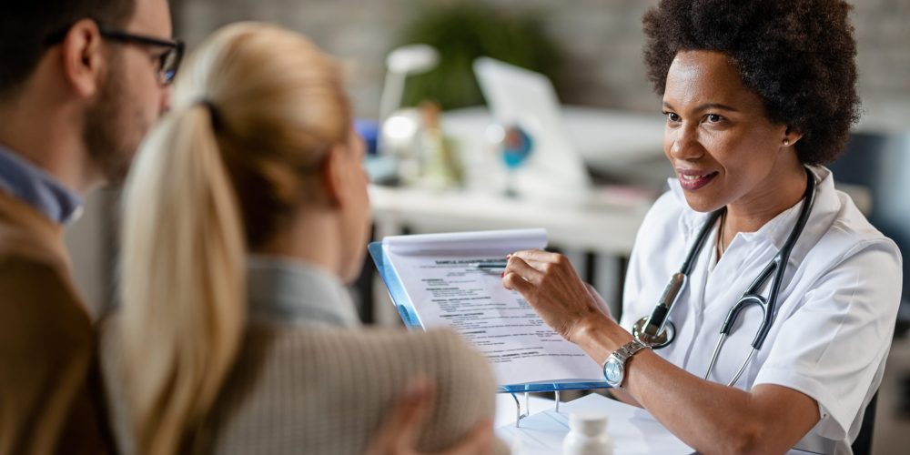 Smiling black female doctor talking to a couple while analyzing their medical reports during consultations at clinic.