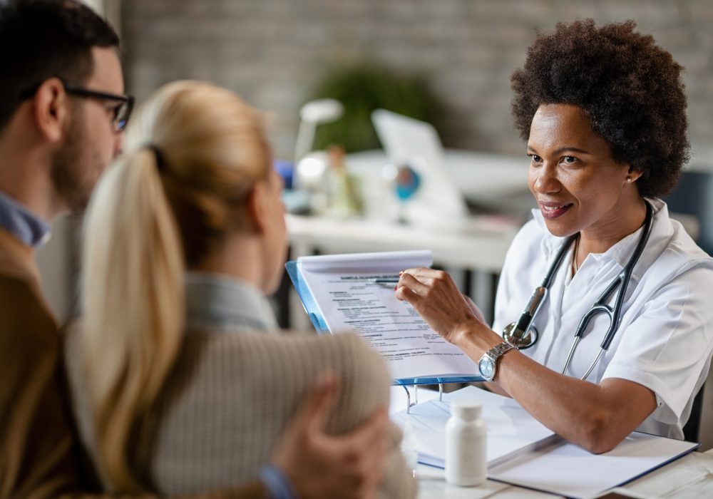 Smiling black female doctor talking to a couple while analyzing their medical reports during consultations at clinic.